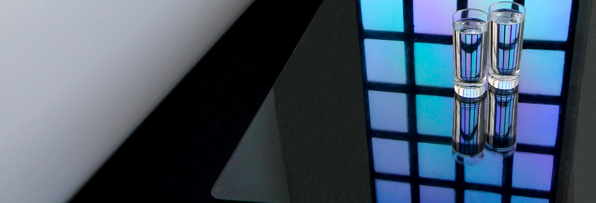 two clear glasses filled almost to the top with fresh water are sitting on top of a black table. on the table and through the water in the glasses, there is a reflection of a stain glass window with square blue and purple ombre tiles.